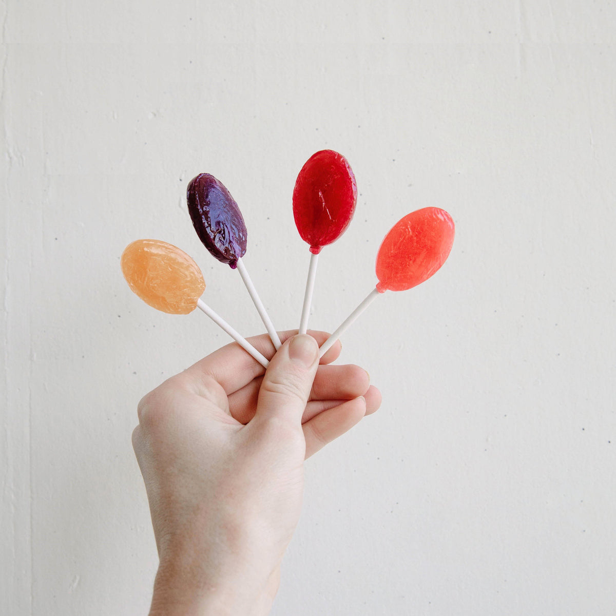 A hand holding four colorful lollipops with orange, purple, red, and pink candies against a plain white background.