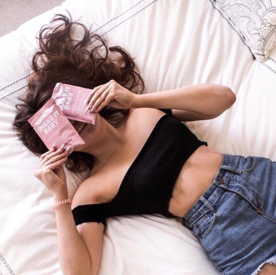 Woman lying on a bed, wearing a black off-shoulder top and blue jeans. Her hair is spread out, and she's holding two pink packets labeled "SHAKE IT BABY" over her eyes. Pillows and white bedding are visible in the background.