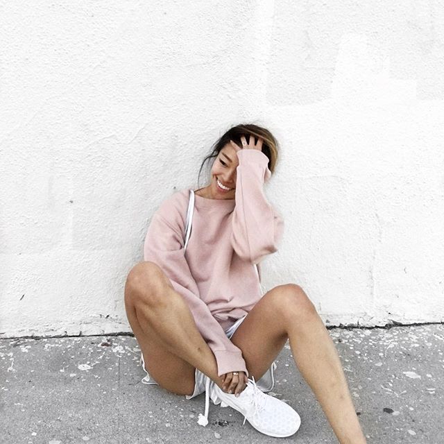 A person sitting against a white wall, smiling and touching their hair. They are wearing a pink sweatshirt, white shorts, and white sneakers. The concrete ground shows some chipped paint.