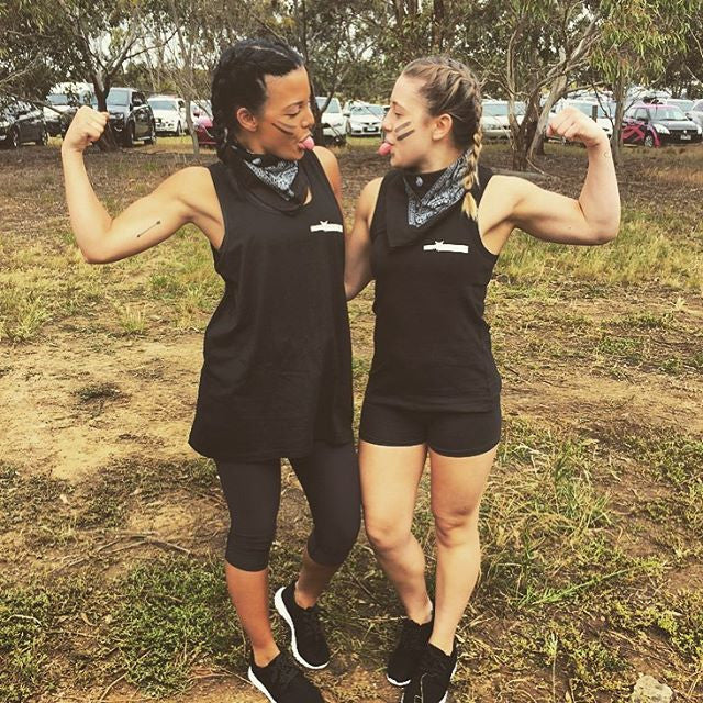 Two women in matching black athletic outfits and bandanas stand outdoors on grassy ground. They face each other, flexing their arms and playfully sticking out their tongues. Cars and trees are visible in the background.