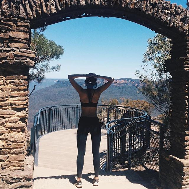A person wearing athletic clothing and a cap stands under a stone archway, overlooking a scenic mountain view with clear skies. Trees frame the pathway leading to a railing that extends out over the landscape.