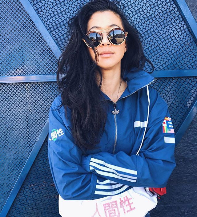A person with long black hair wearing round sunglasses and a blue Adidas jacket stands with arms crossed. The background features a textured metal surface.