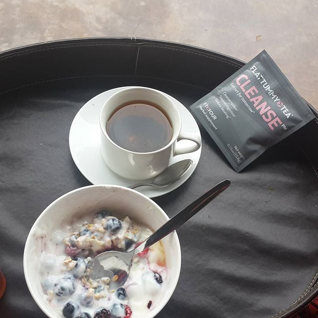 A bowl of yogurt with granola and berries sits beside a cup of tea on a black tray. A packet labeled "Flat Tummy Tea CLEANSE" is nearby.