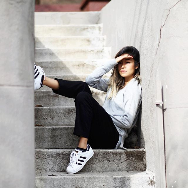 A person sits on concrete stairs, leaning against a wall. They wear a gray sweatshirt, black pants, and white sneakers. Their right hand shields their eyes from the sunlight, and their left leg is bent up.