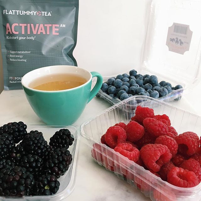 A green mug filled with tea is surrounded by open containers of blackberries, raspberries, and blueberries. A package labeled "Flattummy Tea Activate AM" is seen in the background.