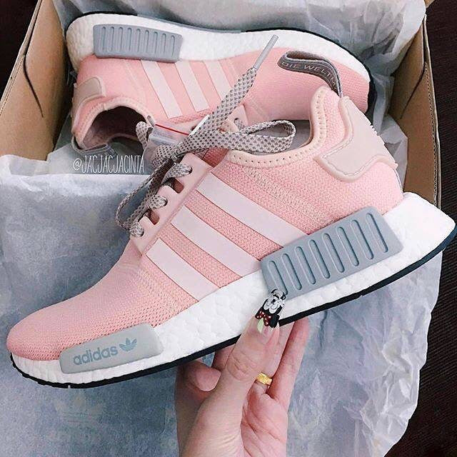 A pair of pink Adidas sneakers with white stripes and gray accents is displayed inside a shoebox. There is a decorative lace charm on one of the shoes. The sneakers rest on white tissue paper.