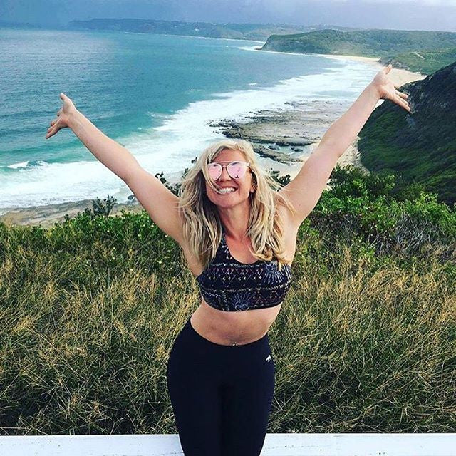A woman with long blonde hair wearing sunglasses and a sports bra poses with arms raised and smiles widely. She stands in front of a scenic coastline with cliffs, green hills, and a blue ocean under a cloudy sky.