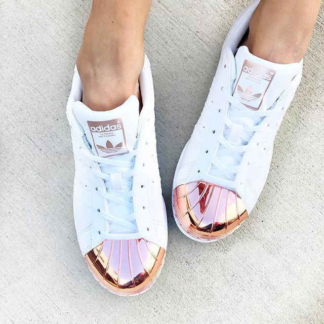 A pair of white Adidas sneakers with metallic rose gold toe caps on a light concrete surface. The shoes have white laces and the Adidas logo on the tongue. The wearer is standing, and only their legs and feet are visible.