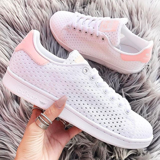 A pair of white sneakers with mesh texture and light pink accents on the heel and tongue, held by a hand with manicured nails and rings. The sneakers are placed on a fluffy gray surface.