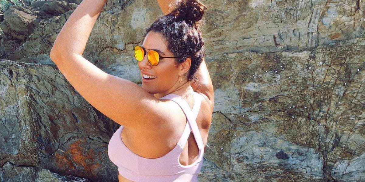 A woman wearing sunglasses and a light purple sports bra smiles while posing outdoors in front of a large rock formation.