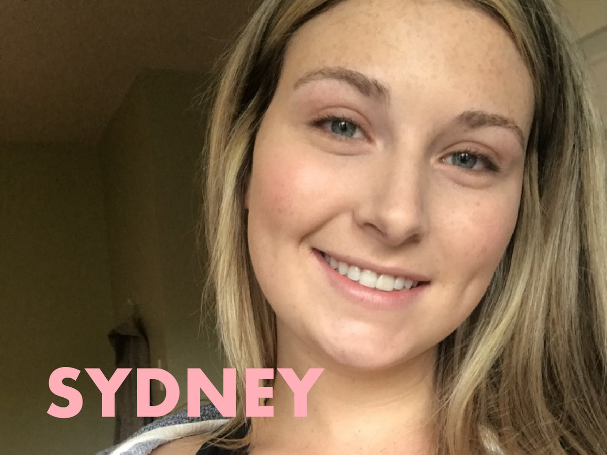 A person with long hair smiles warmly at the camera. The background is softly blurred. The word "SYDNEY" is written in large pink letters at the bottom left corner.