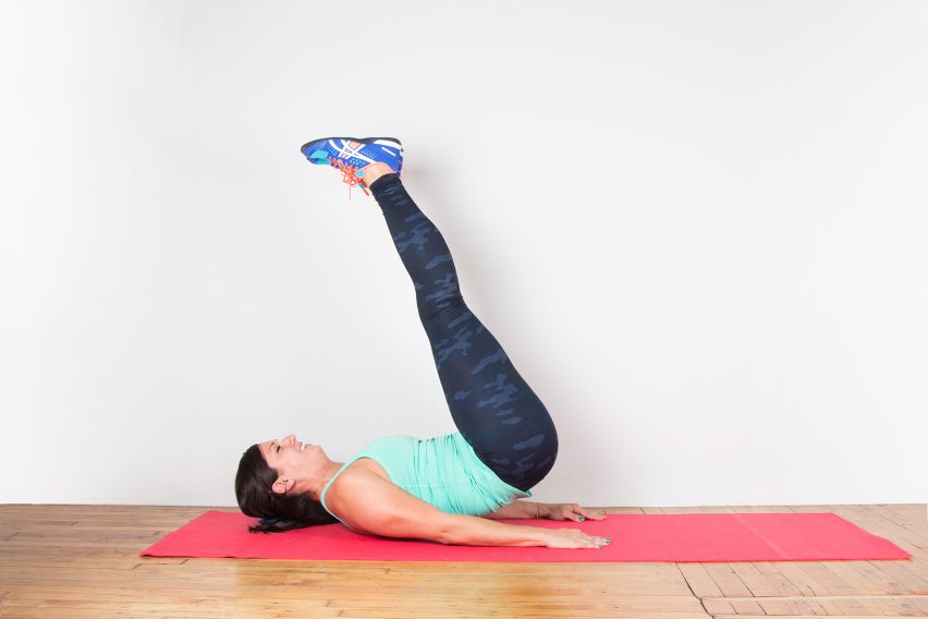 A person in athletic wear is performing a leg raise exercise on a red yoga mat. They are lying on their back with legs extended upwards and arms resting at their sides. The background is a plain white wall and a wooden floor.