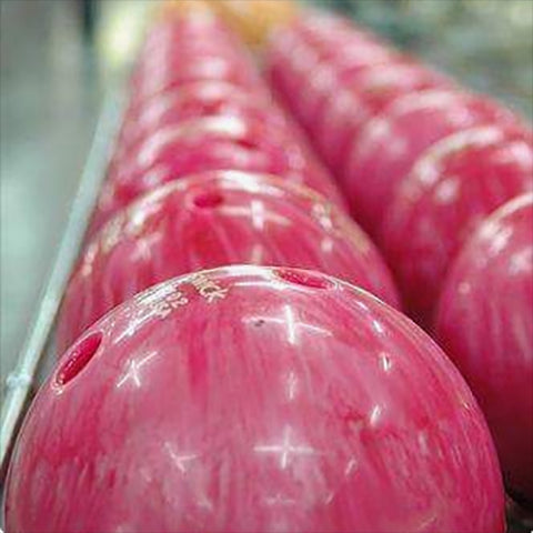 A row of pink bowling balls with finger holes are neatly lined up on a rack. The balls are slightly reflective, showing some light reflections, and are situated in what appears to be a bowling alley.