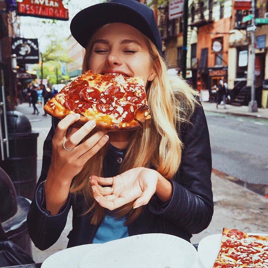 Person wearing a black cap and jacket, happily eating a large slice of pizza with pepperoni toppings at an outdoor cafe. Urban street scene in the background with buildings and signs.