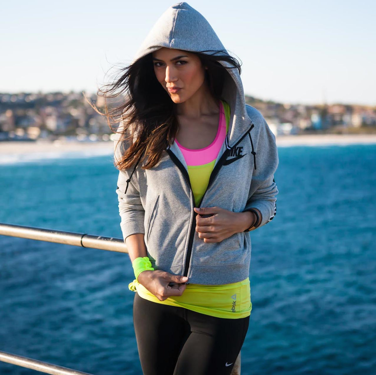 Woman in a gray hooded sweatshirt and neon athletic wear stands on a pier by the ocean. Her hair is blowing in the wind, and a coastal town is visible in the background.