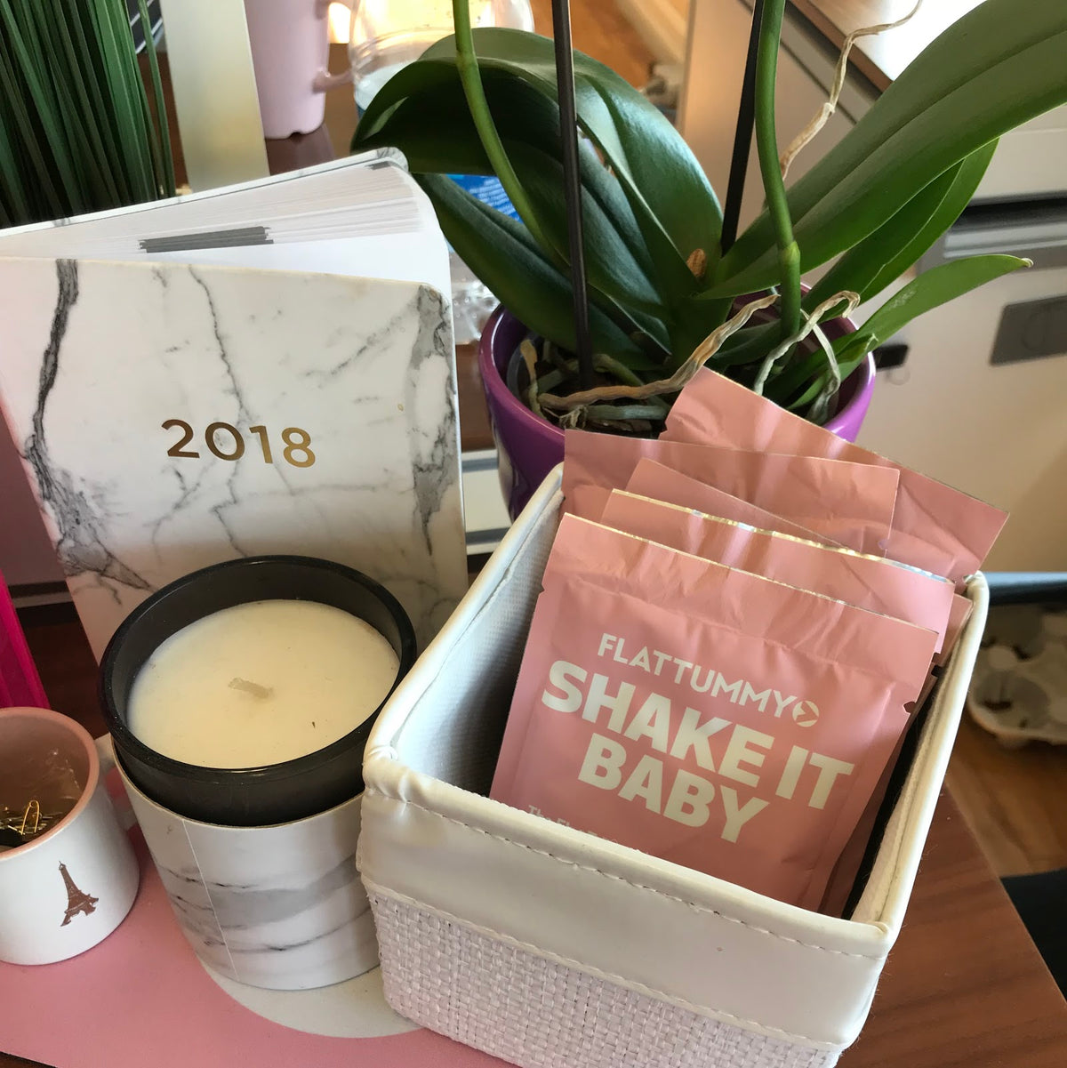A desk scene with a 2018 marble-patterned planner, a candle, an orchid plant in a purple pot, and packets of "Flat Tummy Shake It Baby" in a white basket. A small Eiffel Tower figurine is also visible.