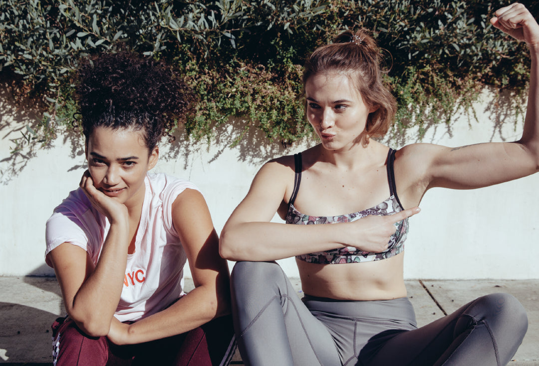 Two young women sit outdoors in athletic wear; one smiles and flexes her arm, pointing at her bicep, while the other rests her chin on her hand and looks at the camera. Green foliage and a white wall are behind them.