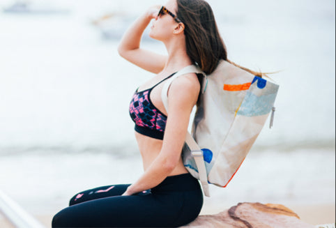 A woman sits on a rock by the beach. She is wearing sunglasses, a sports bra, and leggings, with a large tote bag over her shoulder. The background is the ocean, and the scene conveys a relaxed, summery atmosphere.