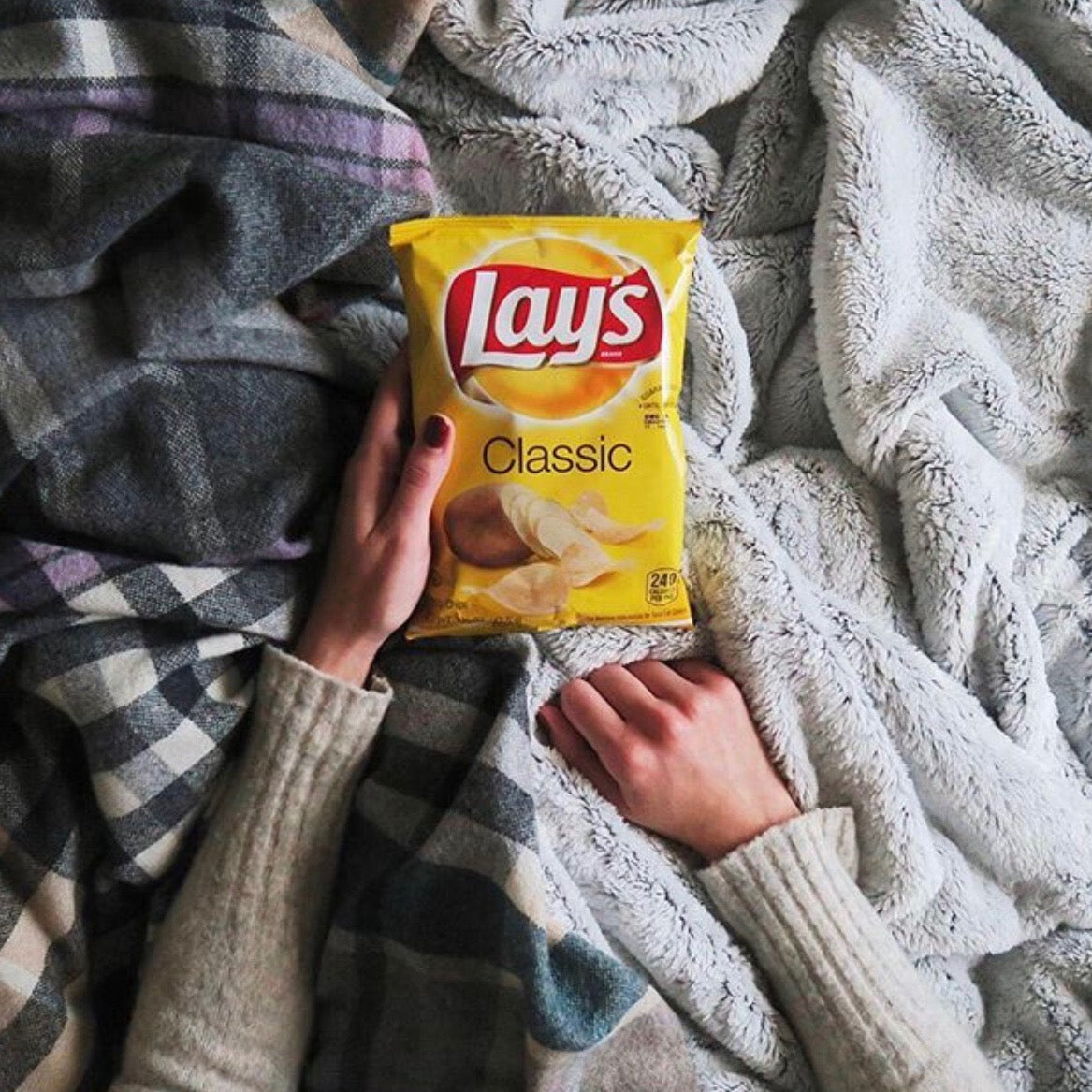 A person in a cozy sweater lies on a soft, patterned blanket, holding a bag of Lay's Classic potato chips. Their hands are visible, resting on the blanket, creating a relaxed and comfortable scene.