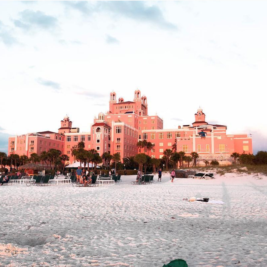 A large pink hotel with multiple towers stands by a sandy beach. Palm trees line the area, and people are gathered on the sand near lounge chairs. The sky is clear with a hint of sunset.