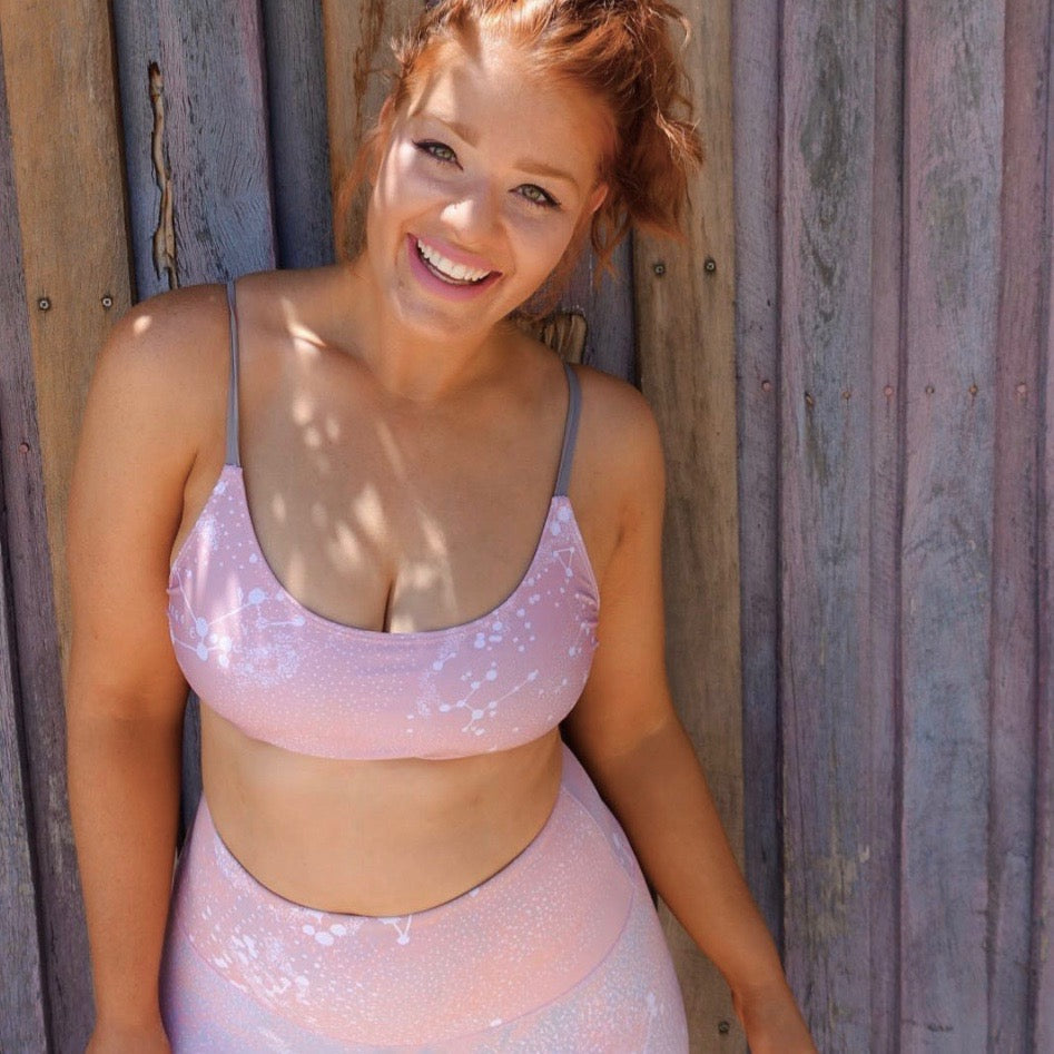 A person with red hair smiles while wearing a pink and white patterned bikini against a wooden and corrugated metal background in the sun.