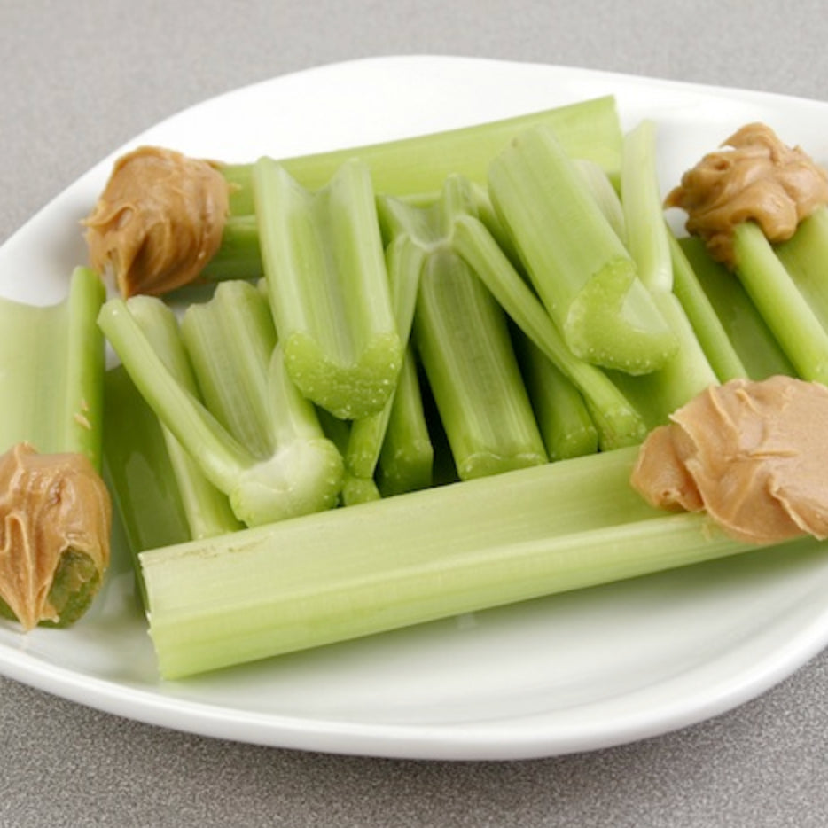 A white plate with several celery sticks topped with dollops of peanut butter on a gray surface.