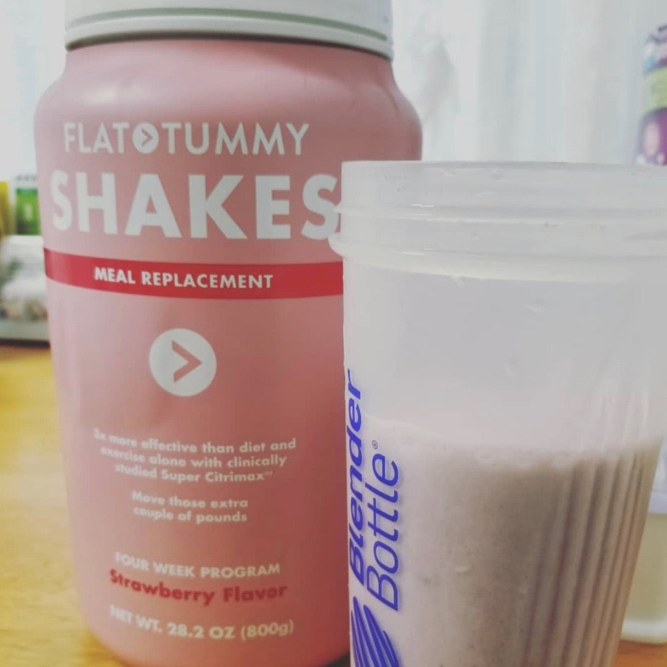 A container of Flat Tummy Shakes meal replacement powder sits next to a clear Blender Bottle filled with a light pink shake on a table.