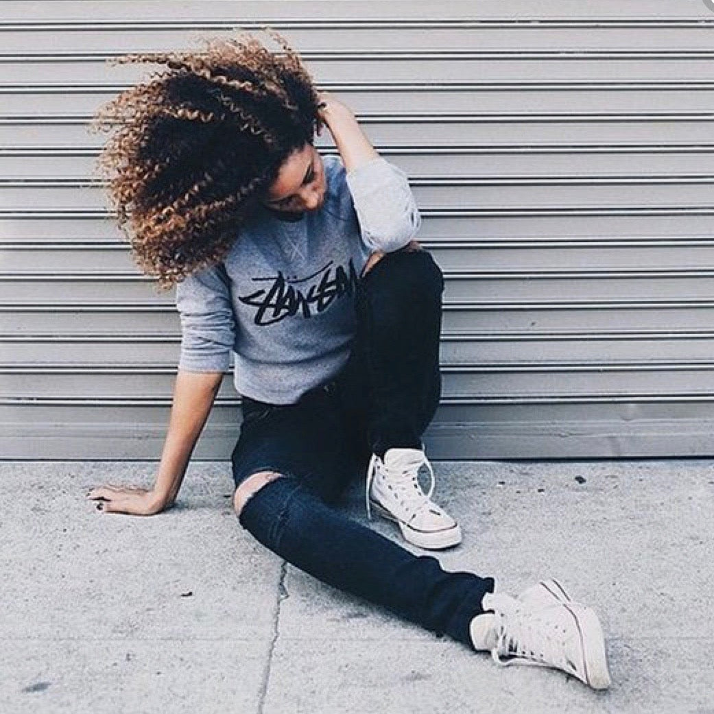 A person with curly hair sits on the pavement in front of a closed garage door. They wear a gray sweatshirt, ripped black jeans, and white sneakers, with one knee bent and one hand touching their hair.