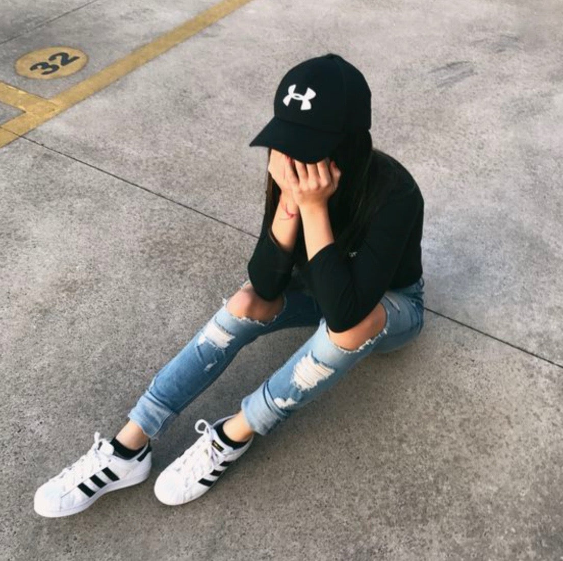 Person sitting on the ground with their head in their hands. They're wearing a black cap, black long sleeve shirt, ripped blue jeans, and white sneakers with black stripes. The ground is concrete with a painted number 32 visible.