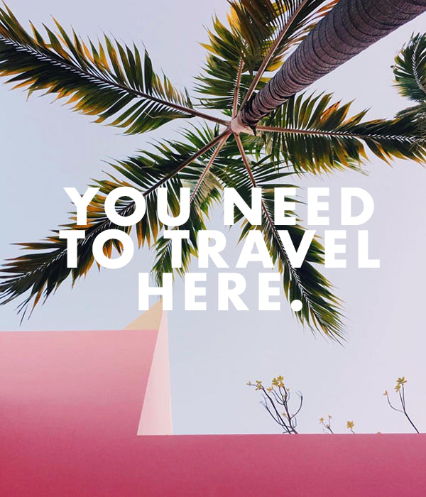 A tall palm tree stretches over a pink building against a clear sky. White text in the foreground reads, "YOU NEED TO TRAVEL HERE." Small branches with few leaves peek over the building edge.