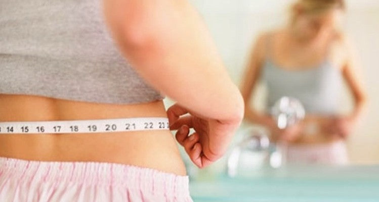 A person in a gray shirt and pink shorts is measuring their waist with a tape measure in front of a mirror. They are pinching the tape to check the measurement, and their reflection is visible, slightly out of focus, in the background.