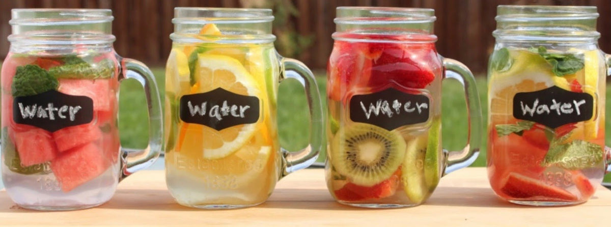 Four mason jars filled with water and various fruit slices, including watermelon, lemon, kiwi, strawberry, and mint, are lined up on a wooden surface. Each jar has a chalkboard label with the word "Water." A blurred background of grass is visible.