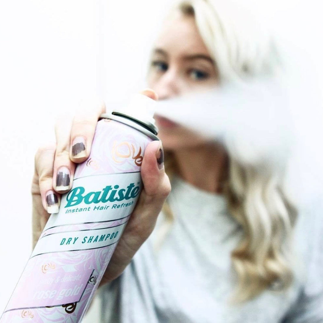 A woman with long blonde hair sprays Batiste dry shampoo, labeled "Rose Gold," from a can. The background is blurred, and her face is slightly out of focus, emphasizing the can she is holding.