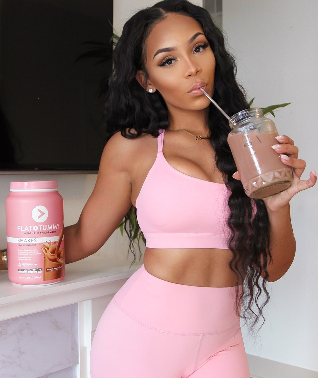 Woman in pink activewear drinking a Flat Tummy Shake, with product container on counter.
