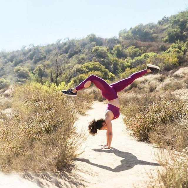 A woman in a pink athletic outfit does a handstand on a sunny dirt trail surrounded by dry bushes and green hills.