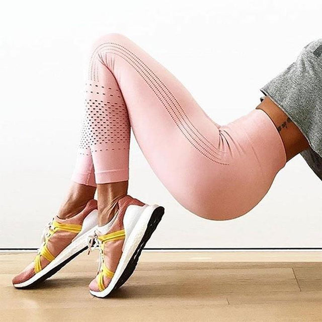 A person in a grey top and pink leggings performs a glute bridge exercise indoors, with their feet flat on the wooden floor and wearing pink and white athletic shoes.