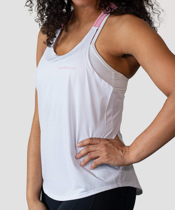 A person with curly hair is wearing a Babe Tank by Flat Tummy Co, which is a white athletic tank top featuring pink shoulder straps. Their hand is resting on their hip, and the background is plain white. The word "FLATTUMMY" can be seen on the breathable, quick-dry tank top.