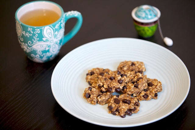 A light blue plate with several oatmeal chocolate chip cookies placed on a dark wooden table. Next to the plate is a teal mug with white floral designs containing tea and an adjacent tea infuser.