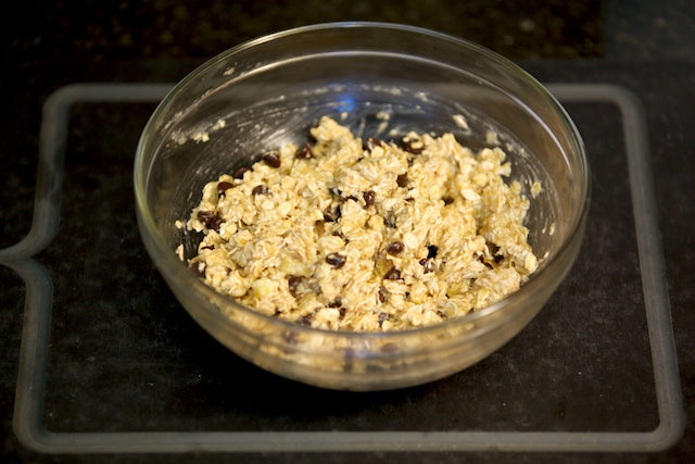 A glass bowl filled with a mixture of oats, chocolate chips, and other ingredients sits on a dark countertop. The mixture looks partially blended, suggesting the initial stages of preparing a recipe.