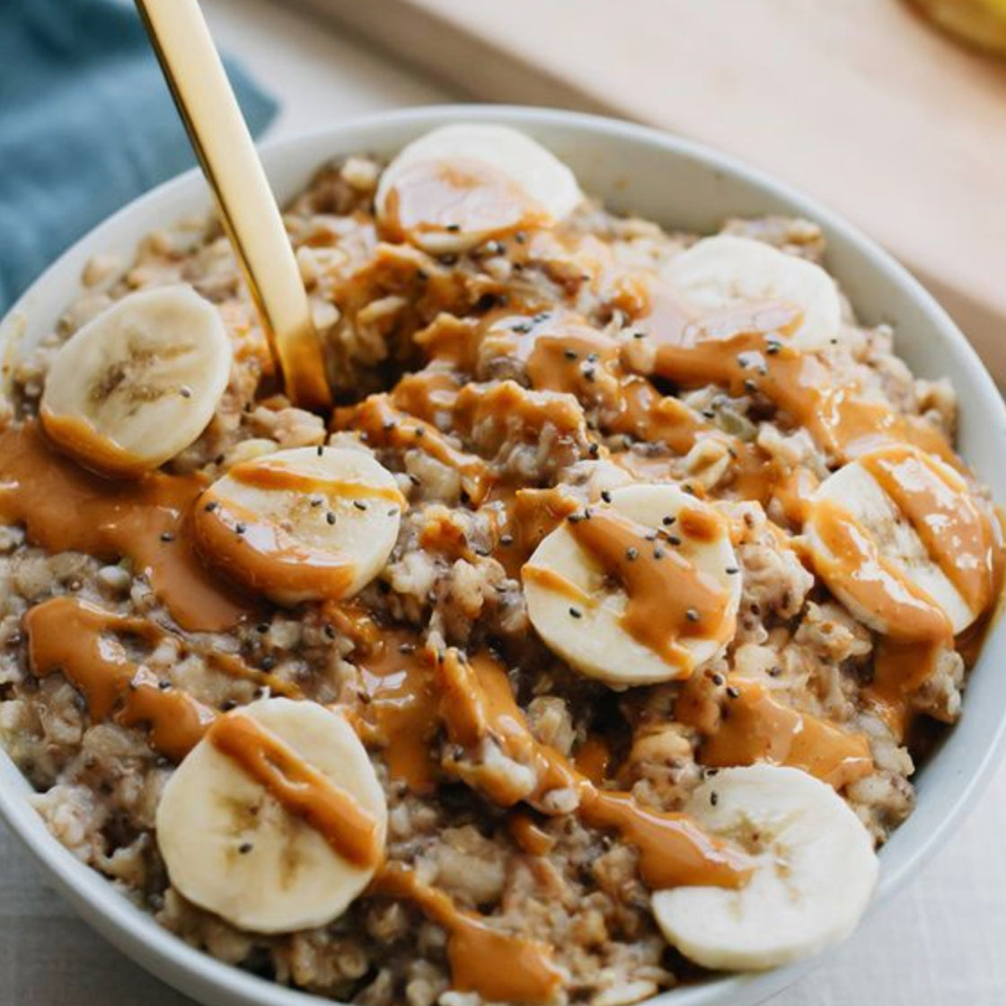 A bowl of oatmeal topped with banana slices, a drizzle of peanut butter, and sprinkled chia seeds. A gold spoon is placed in the bowl. A blue napkin and a wooden board are partially visible in the background.