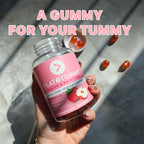 A hand holding a bottle of Flat Tummy Co Apple Cider Vinegar Gummies. The pink-labeled bottle sits against a textured background with a few gummies scattered around. Above, the text reads "A GUMMY FOR YOUR TUMMY," emphasizing the benefits for gut health.