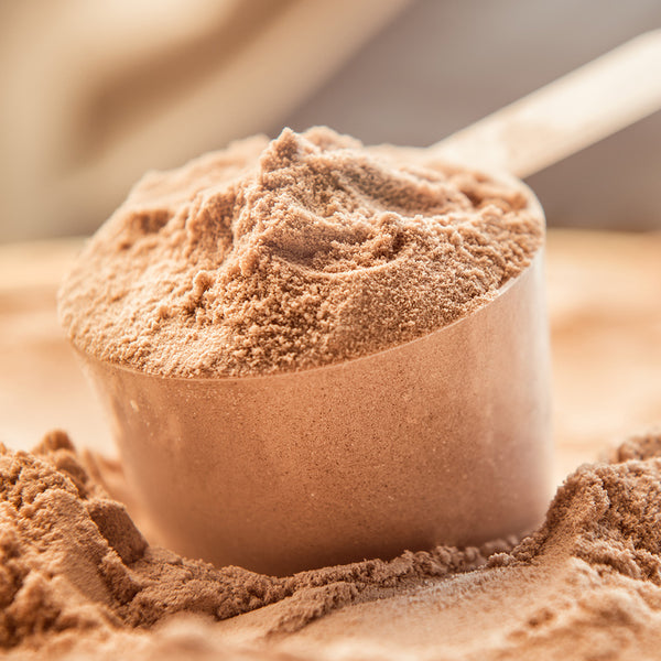 A close-up of a scoop filled with chocolate protein powder. The scoop is nestled in a pile of more powder, creating a textured, fine-grained surface. The background is softly blurred, highlighting the fine detail of the powder in the scoop.